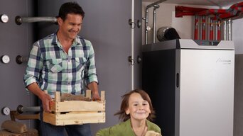 Ein lächelnder Mann und ein fröhliches Kind tragen gemeinsam eine Holzkiste in einen Heizraum, in dem eine moderne bösch Heizungsanlage steht. | © Walter Bösch GmbH & Co KG