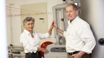 Ein älteres Paar steht neben einer Heizungsanlage in einem Heizraum. Sie lachen und berühren sich spielerisch mit den Händen in einem "High Five"-Stil. Die Frau trägt eine weiße Bluse und einen dunklen Rock, der Mann ein weißes Hemd und dunkle Hose. Im Hintergrund sind isolierte Rohre und technische Ausrüstung sichtbar. | © bösch heizung.klima.lüftung