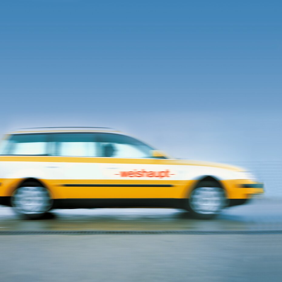Verschwommenes Bild eines schnell fahrenden gelb-weißen Autos mit dem Weishaupt-Logo auf der Seite, vor einem blauen Himmel.