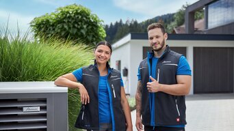 Zwei lächelnde Techniker, eine Frau und ein Mann, stehen neben einer Außeneinheit einer Wärmepumpe. Beide tragen Firmenkleidung mit dem Logo von bösch. Im Hintergrund ist ein modernes Wohnhaus sichtbar. Der Techniker hält einen Daumen hoch. | © bösch heizung.klima.lüftung