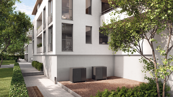 Moderne Wohnanlage mit weißen Fassaden und mehreren Fenstern. Vor dem Gebäude befindet sich ein gepflegter Grünstreifen mit Bäumen und Sträuchern. Im Vordergrund sind zwei MOZART Wärmepumpen sichtbar.