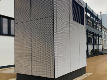 Hohes, modernes Lüftungsgerät zur Vorfilterung von Außenluft, ummantelt mit schwarz-weißem Gitter, steht auf einem gepflasterten Boden vor einem mehrstöckigen Gebäude mit großen Fenstern. Das Gerät ist an der Basis mit einem schwarzen Rand versehen und von einer Schicht Kies umgeben. | © Walter Bösch GmbH & Co KG