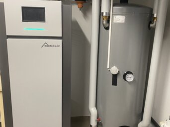 Sole/Wasser-Wärmepumpe KLIMT von bösch in einem Heizungsraum, neben einem großen grauen Warmwasserspeicher. Die Wärmepumpe ist mit einem digitalen Display ausgestattet und verbunden mit mehreren Rohrleitungen, Ventilen und Manometern zur Steuerung der Temperatur und Wasserzirkulation. | © Walter Bösch GmbH & Co KG