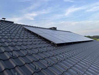 Reihe von dunklen Photovoltaikmodulen auf einem grauen Dachziegel, mit einem leicht bewölkten blauen Himmel im Hintergrund. Die PV-Module sind effizient angeordnet, um maximale Sonneneinstrahlung zu nutzen, und bieten eine umweltfreundliche Energielösung. | © Walter Bösch GmbH & Co KG