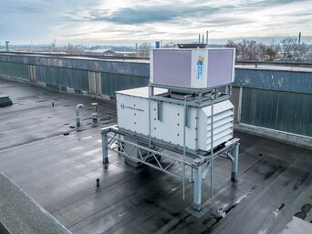 Luftaufnahme eines großen, auf einem Fabrikdach installierten Lüftungssystems. Die Anlage steht auf einer Metallstruktur und ist vorwiegend in Weiß gehalten, mit einem oberen Gitter in Silber. Das Dach ist nass, möglicherweise nach einem Regenschauer. Im Hintergrund ist eine städtische Umgebung mit entfernten Gebäuden und leicht bewölktem Himmel zu erkennen. | © INFRANORM®