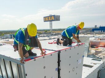 Zwei Montagetechniker auf einem Flachdach montieren sorgfältig eine große metallene RLT-Anlage. Sie tragen Schutzhelme und Sicherheitswesten.