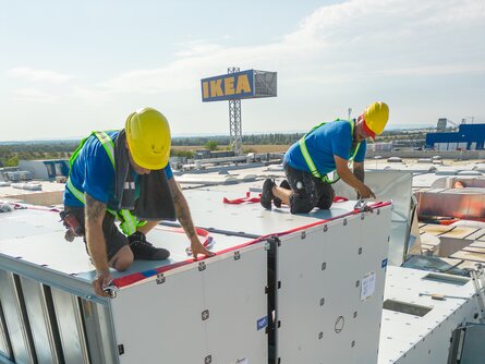 Zwei Montagetechniker auf einem Flachdach montieren sorgfältig eine große metallene RLT-Anlage. Sie tragen Schutzhelme und Sicherheitswesten.