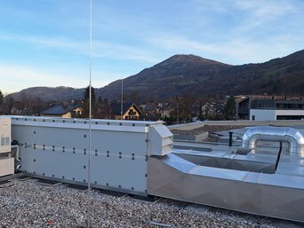 Weitwinkelaufnahme eines Daches bedeckt mit Kies und mehreren Klimaanlagen-Einheiten. Das Dach bietet eine Aussicht auf eine hügelige Landschaft mit Bäumen und einige Gebäude im Hintergrund unter einem klaren Abendhimmel.