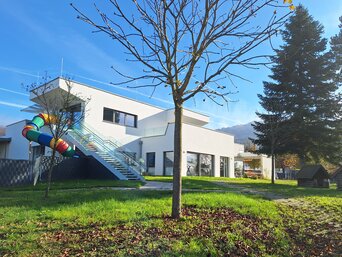Modernes weißes Kindergarten-Gebäude mit bunter Rutsche, die an der Außenseite angebracht ist. Im Vordergrund steht ein einzelner Baum auf einem Rasen mit herbstlich gefärbten Blättern auf dem Boden, unter einem klaren blauen Himmel. | © Marius Projekt ZT GmbH