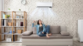 Frau sitzt mit Fernbedienung auf Couch, aus der Klimaanlage kommt frische Luft