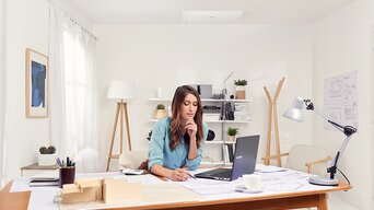 Frau sitzt nachdenkend am Schreibtisch mit Stift in der Hand und Split-Klimaanlage im Hintergrund
